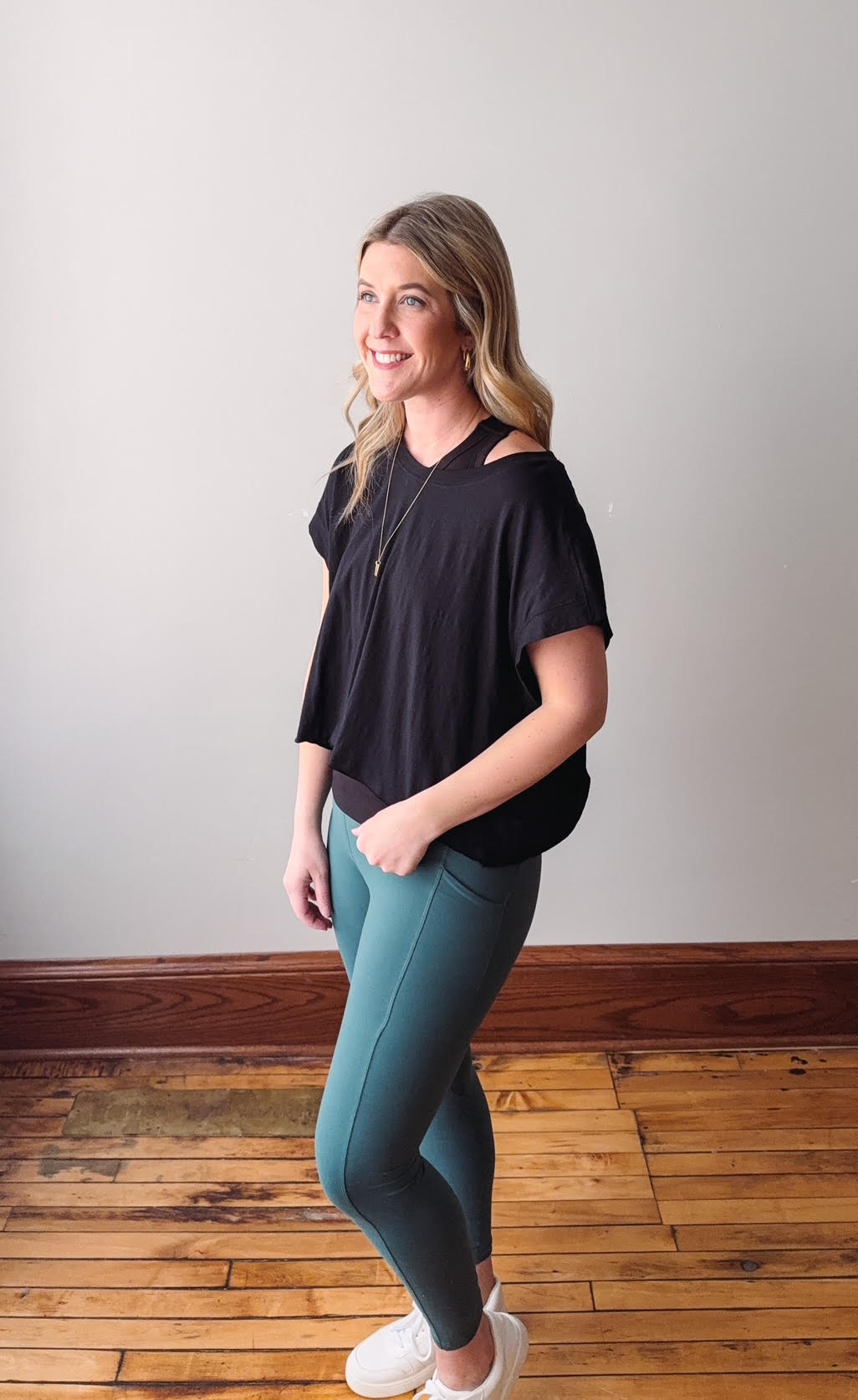 Woman wearing a black top and teal leggings standing on a wooden floor with a white wall background