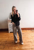 Woman taking a mirror selfie wearing a black shirt and patterned pants in a room with wooden flooring.