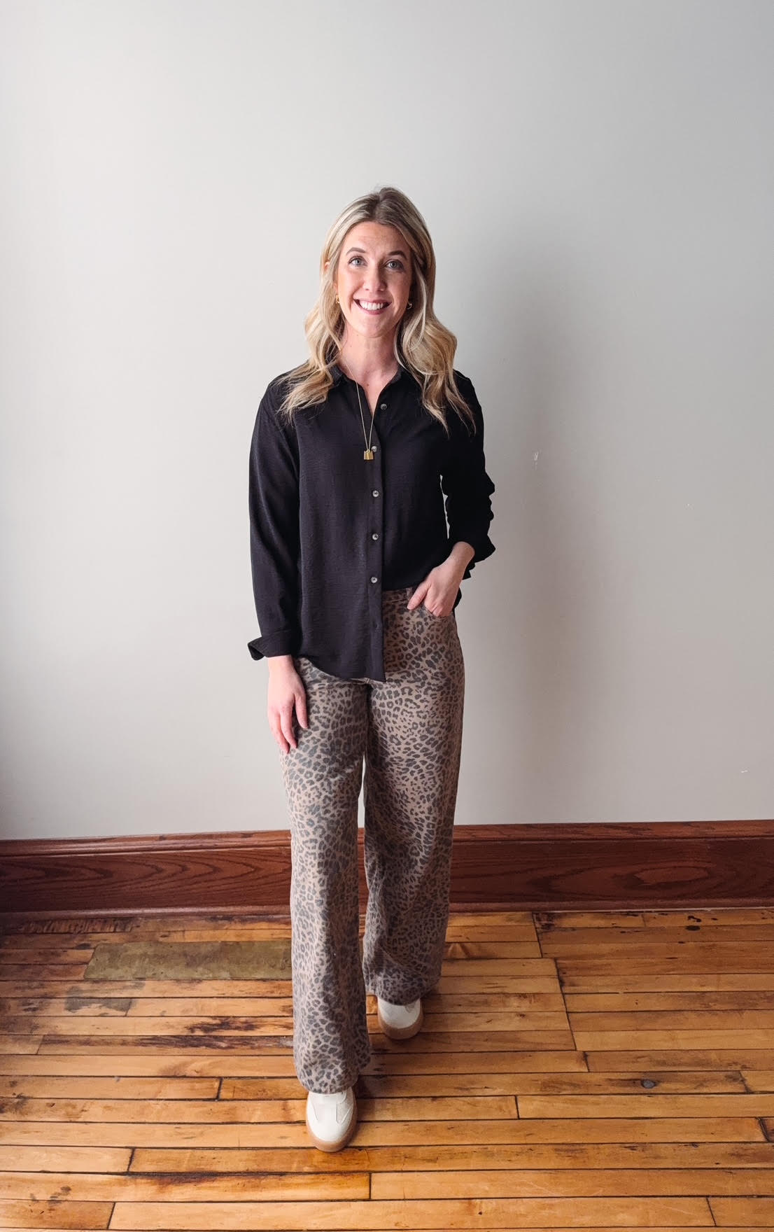 Woman wearing a black shirt and patterned pants standing on a wooden floor against a white wall.