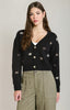 A woman modeling a black knit cardigan with embroidered star and snowflake patterns, featuring gold buttons and a V-neckline, paired with olive green cargo pants against a light gray studio background.