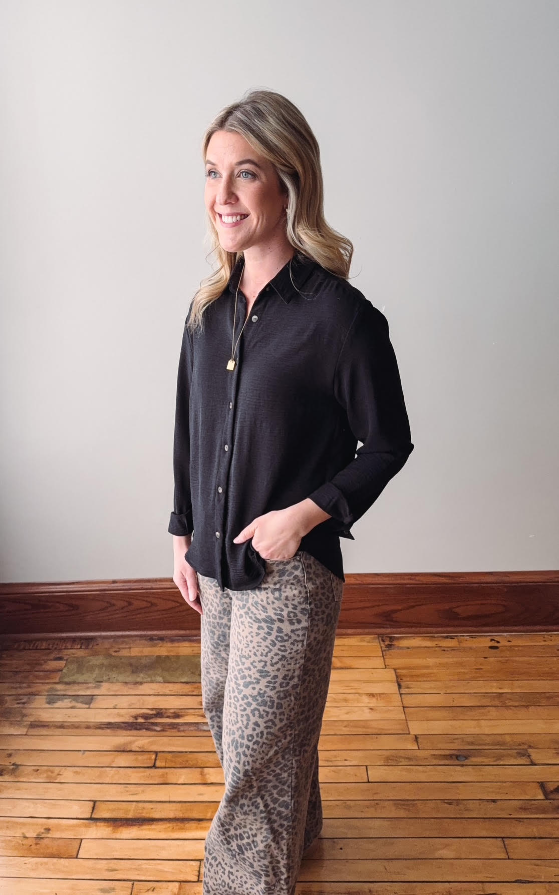 Woman wearing a black shirt and patterned pants standing on a wooden floor with a white wall background