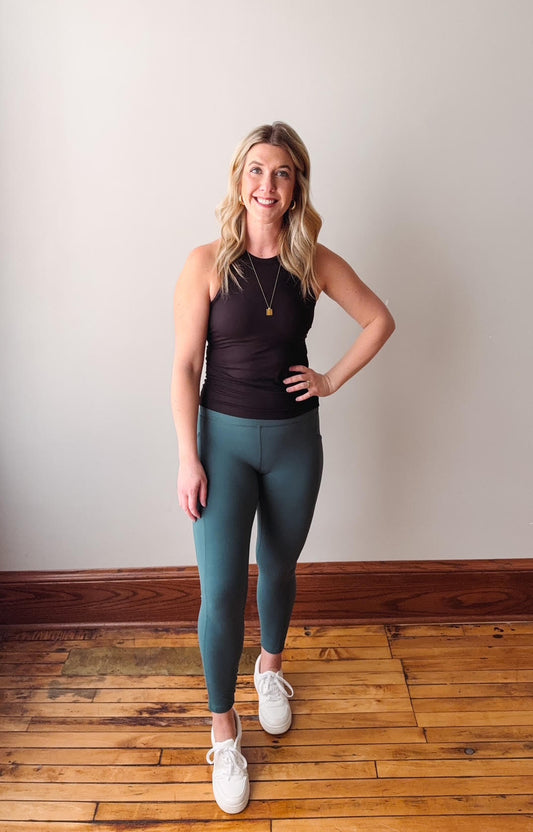 Woman wearing a black tank top and teal leggings standing on a wooden floor.
