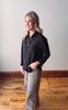Woman wearing a black shirt and patterned pants standing on a wooden floor with a white wall background