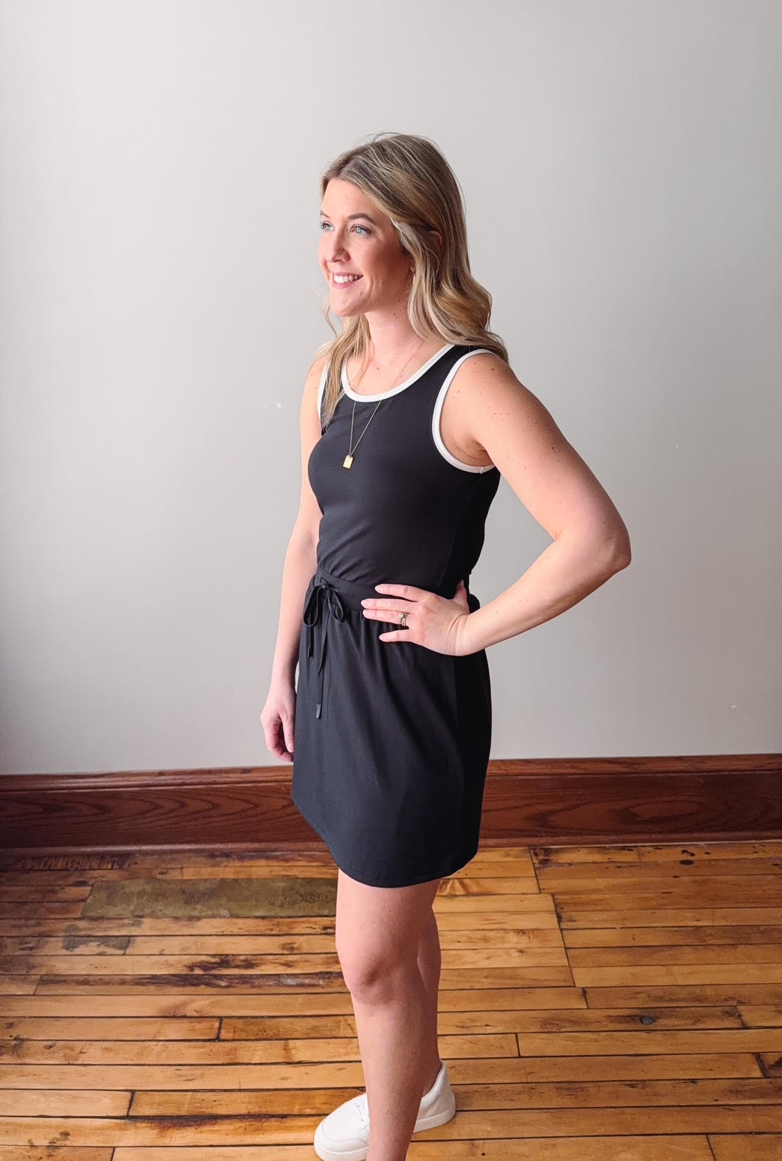 Woman wearing a black dress standing on a wooden floor with a white wall background