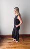 Woman wearing a black sleeveless jumpsuit standing on a wooden floor against a white wall.