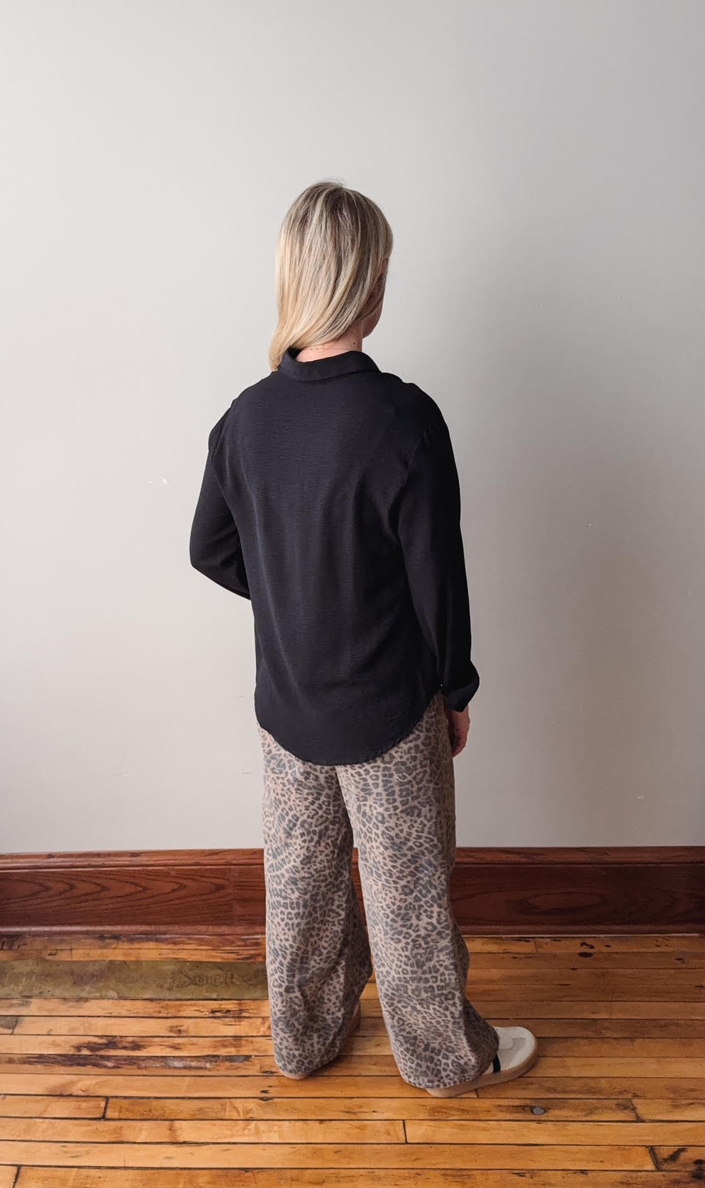 Person wearing a black shirt and patterned pants standing on a wooden floor against a white wall.