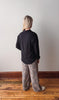Person wearing a black shirt and patterned pants standing on a wooden floor against a white wall.