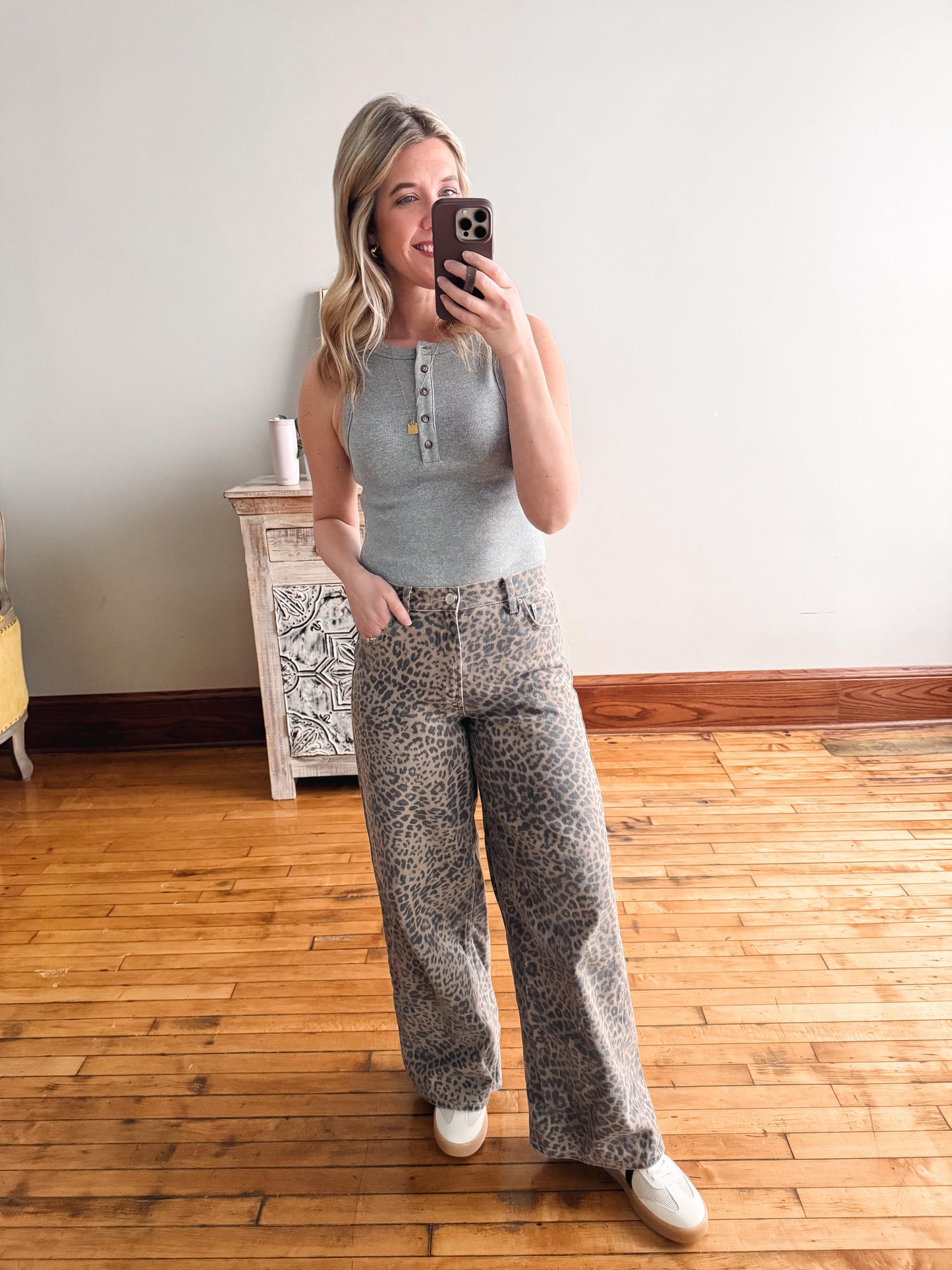 Woman taking a mirror selfie wearing a gray tank top and patterned pants in a room with wooden flooring.