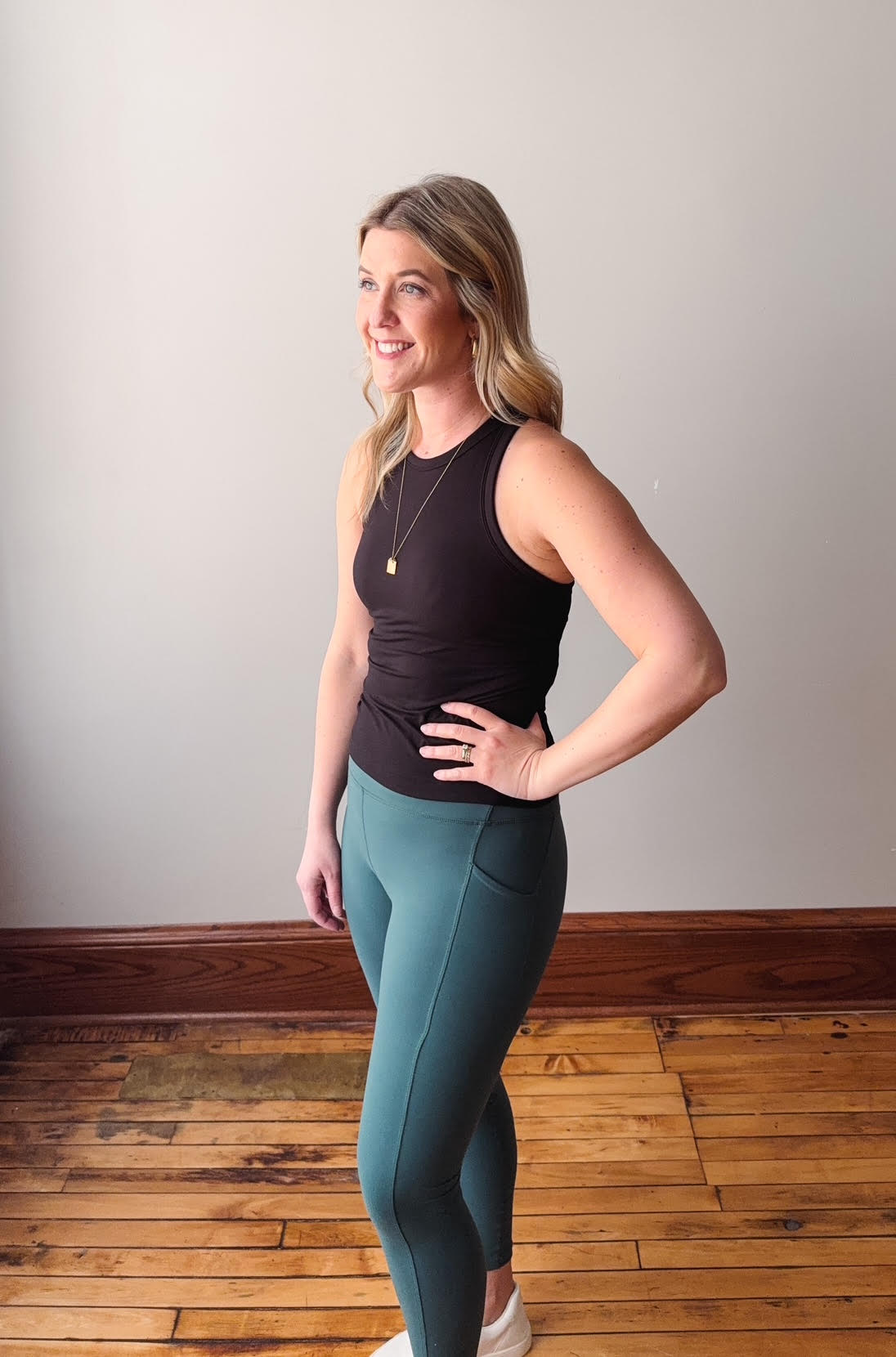 Woman wearing a black sleeveless top and teal leggings standing on a wooden floor.