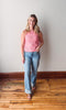 Woman wearing a pink top and blue jeans standing on a wooden floor with a white wall background