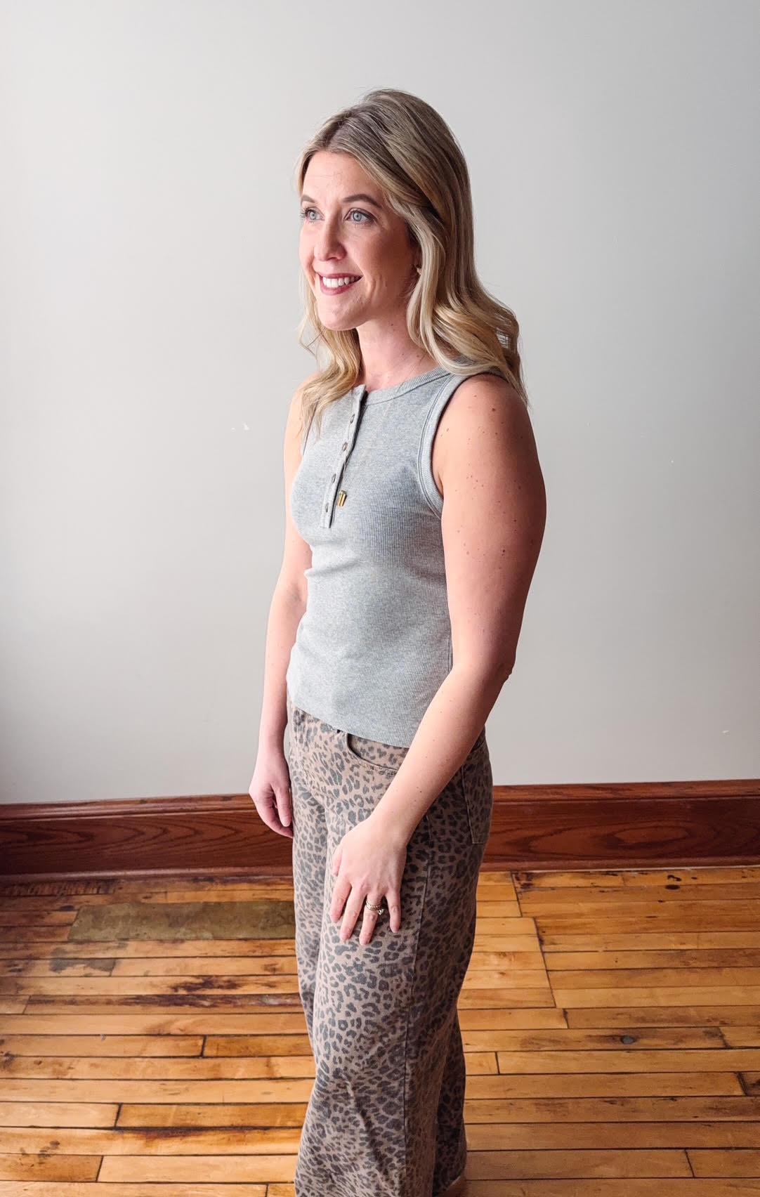 Woman wearing a gray sleeveless top and patterned pants standing on a wooden floor.