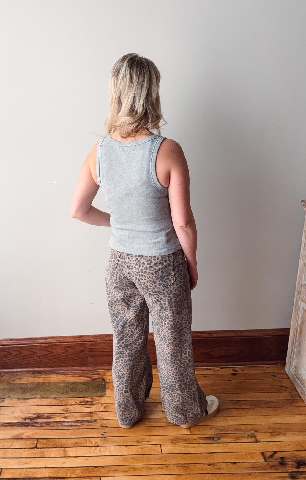 Person wearing a light gray tank top and patterned pants standing in a room with wooden flooring and a white wall.