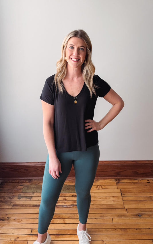 Woman wearing a black top and teal leggings standing on a wooden floor.
