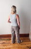Person wearing a light gray tank top and patterned pants standing in a room with wooden flooring and a white wall.