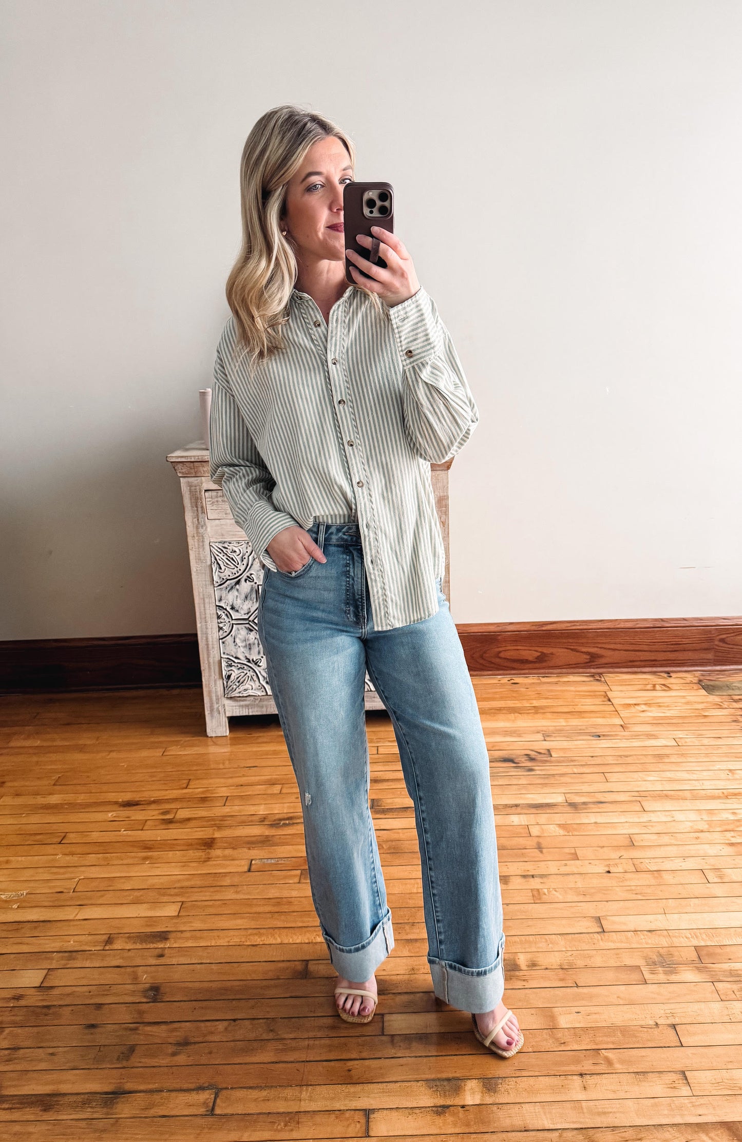 Woman taking a mirror selfie wearing a striped shirt and jeans in a room with wooden flooring.