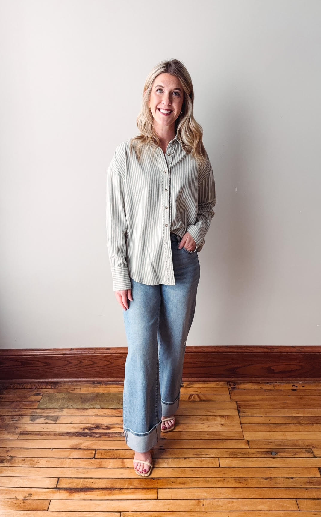 Woman wearing a light-colored shirt and jeans standing on a wooden floor with a white wall background