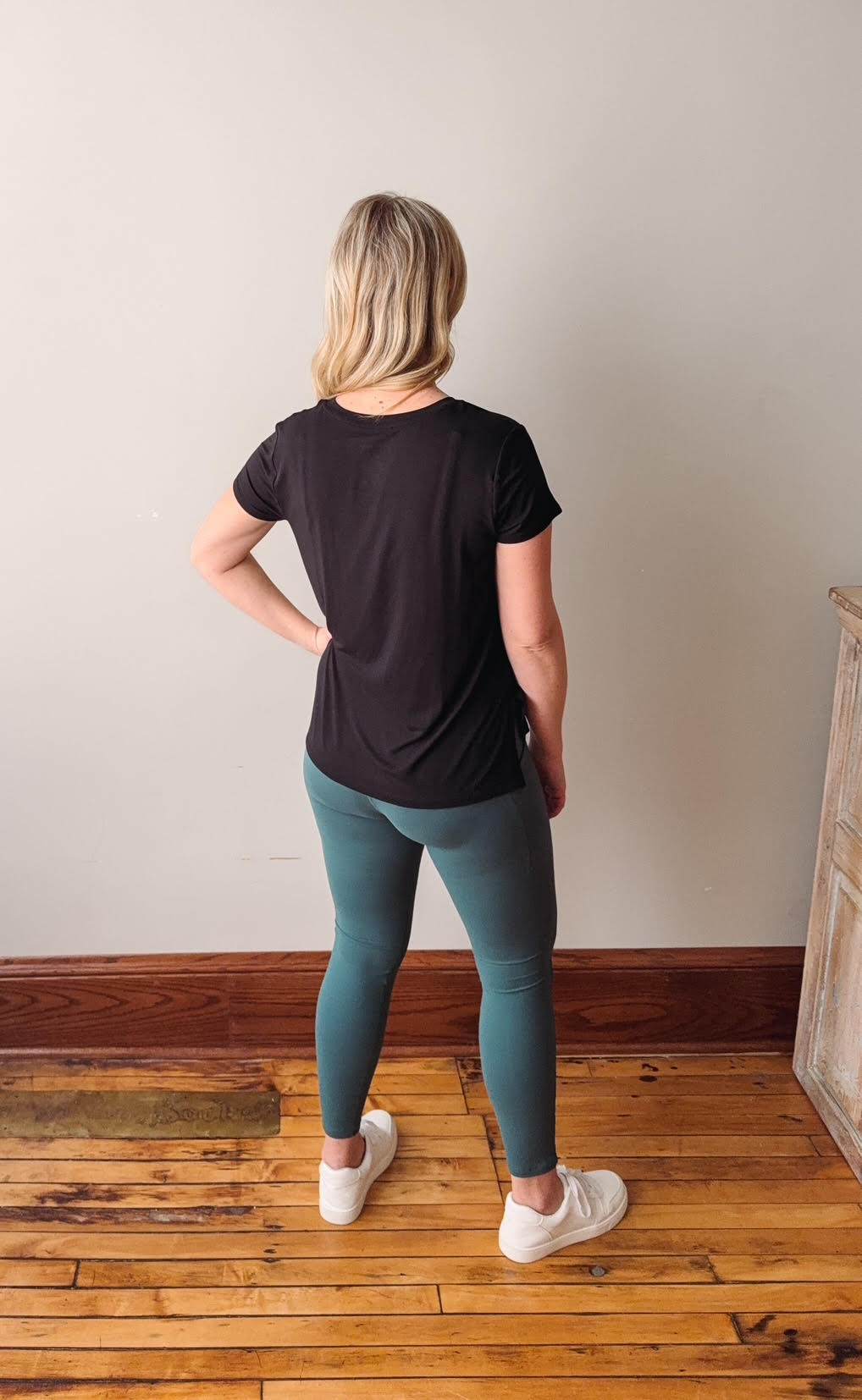 Person wearing a black t-shirt and teal leggings standing on a wooden floor with a plain wall background