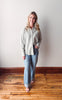 Woman wearing a light-colored shirt and jeans standing on a wooden floor with a white wall background