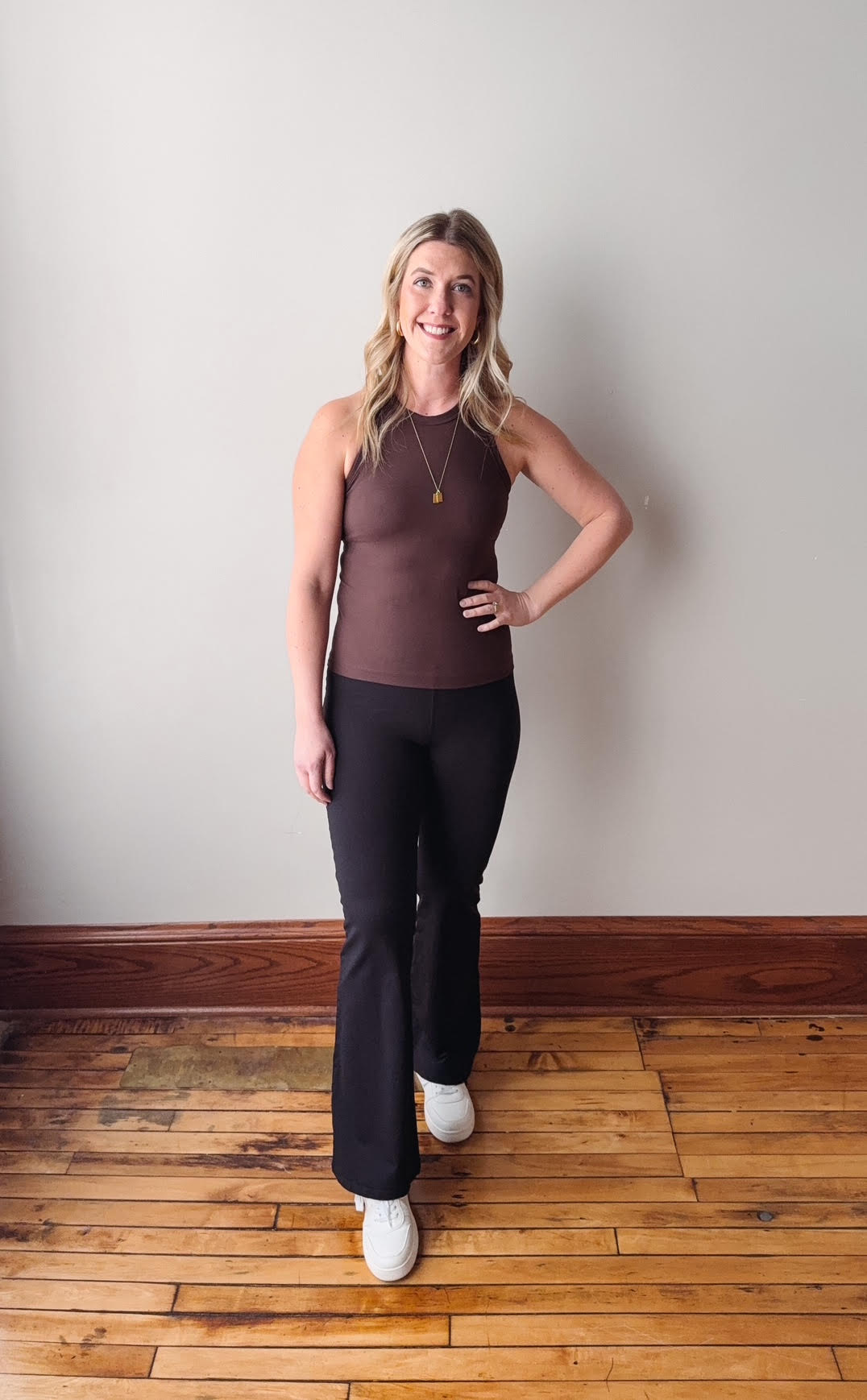 Woman in brown tank top and black leggings standing on a wooden floor with a white wall background