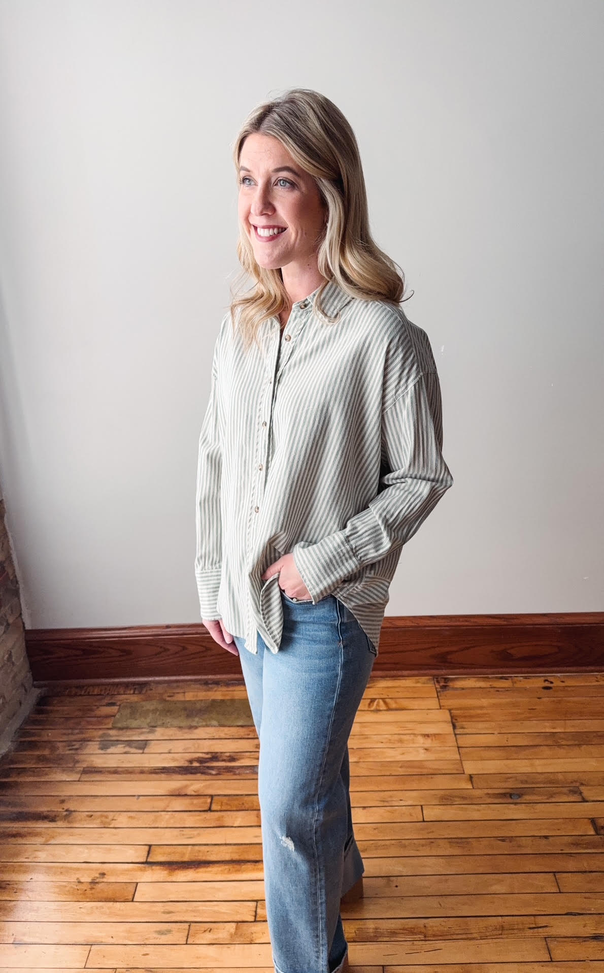 Woman wearing a striped shirt and jeans standing on a wooden floor with a white wall background