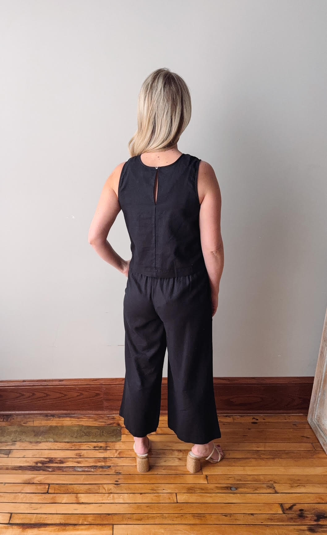 Woman wearing a black sleeveless top and wide-leg pants standing on a wooden floor against a plain wall.