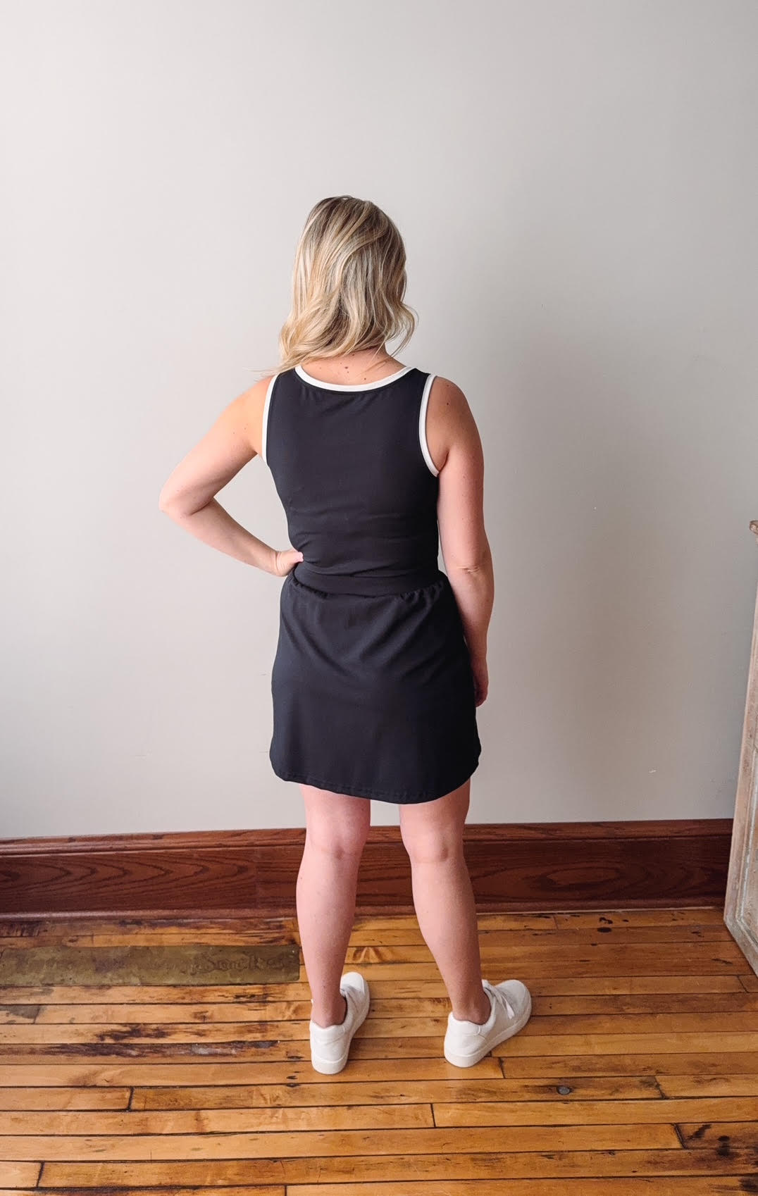 Person wearing a black sleeveless dress standing in a room with wooden flooring and a white wall.
