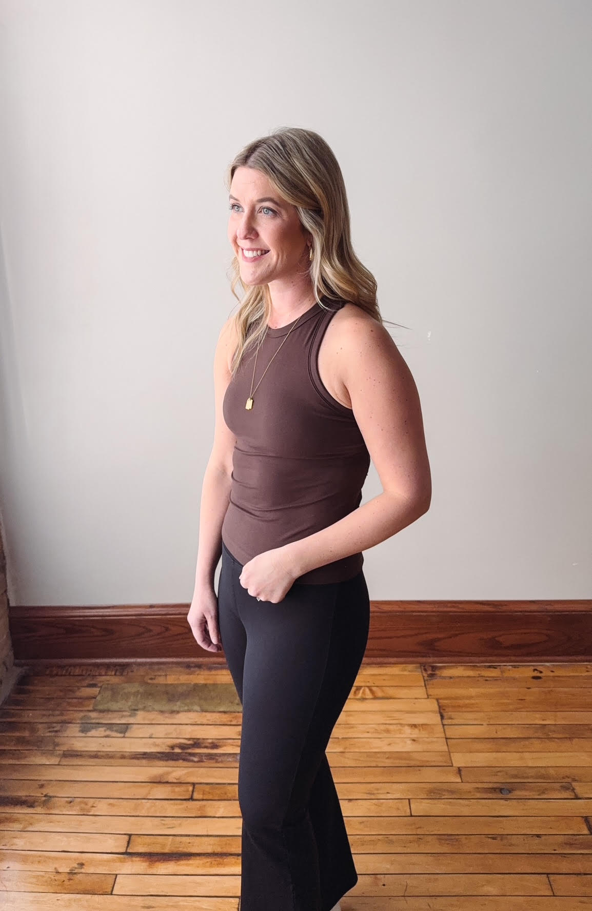 Woman wearing a brown sleeveless top and black pants standing in a room with wooden flooring and a white wall.