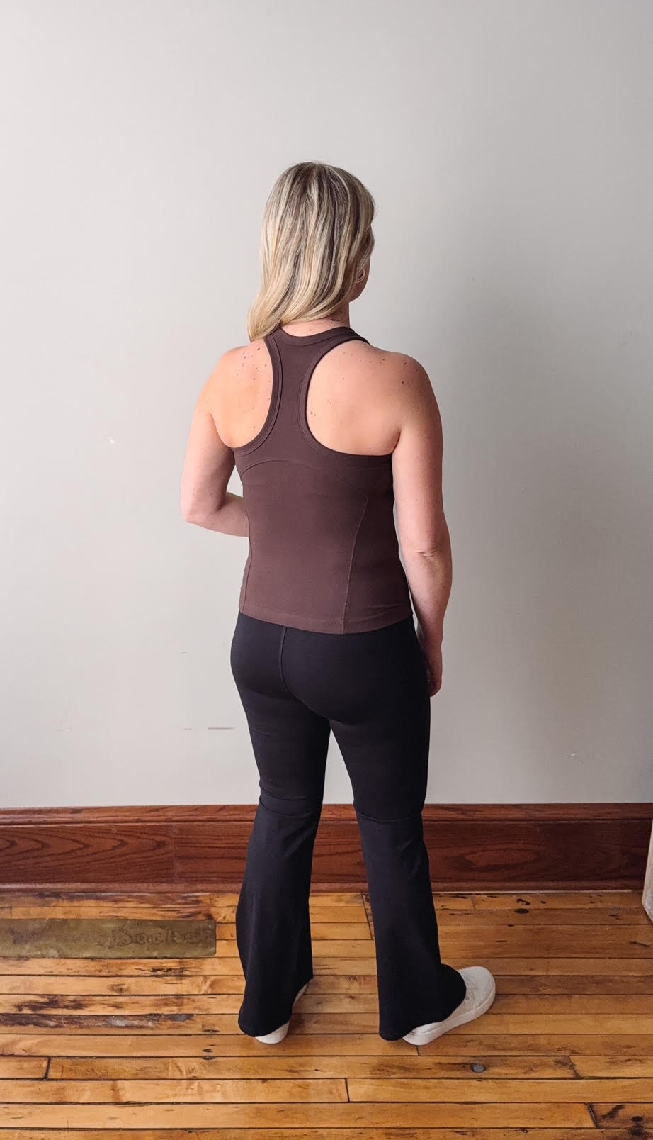 Person wearing a brown tank top and black pants standing on a wooden floor with a white wall background