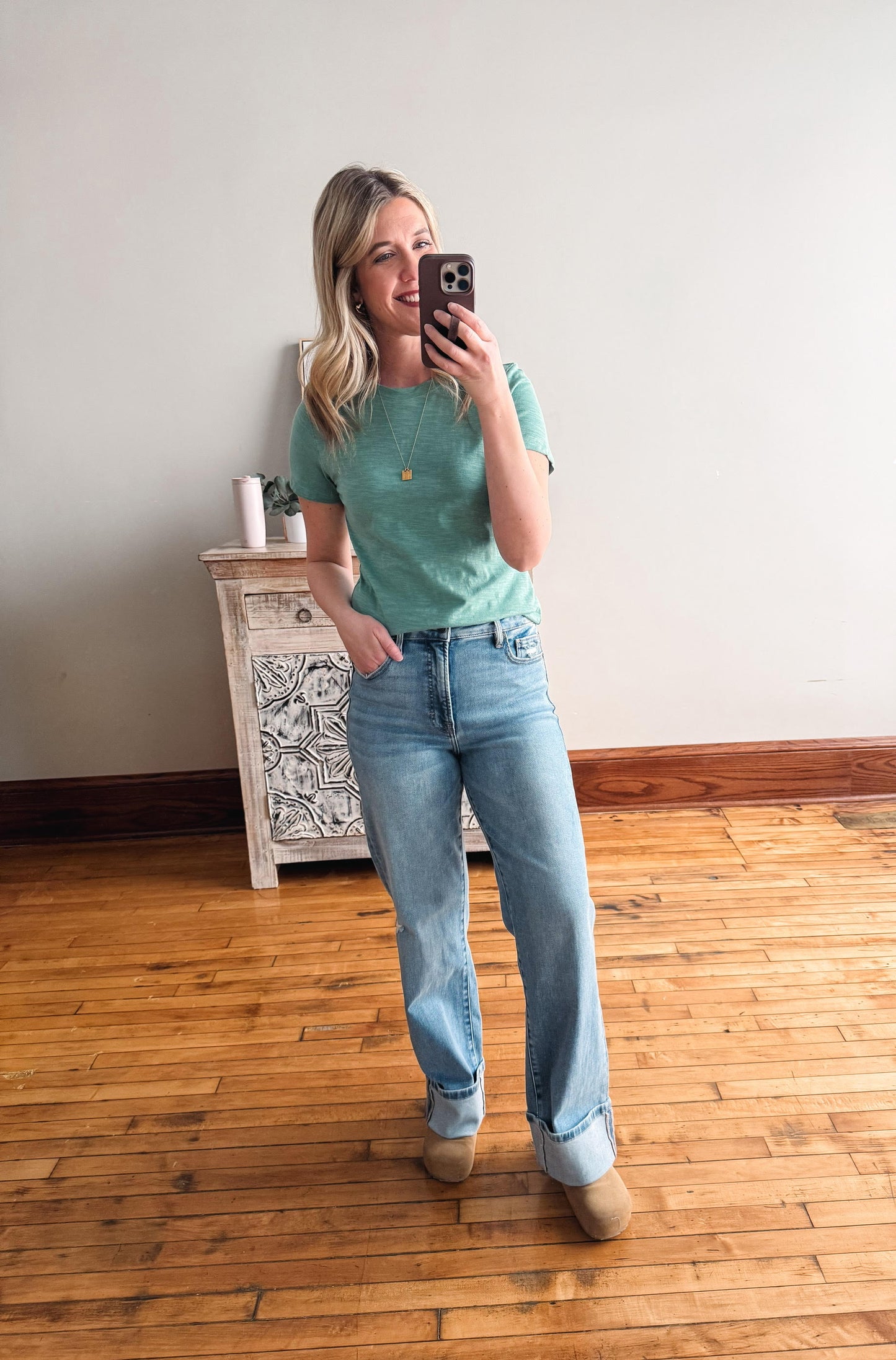 Woman taking a mirror selfie wearing a green t-shirt and blue jeans in a room with wooden flooring.