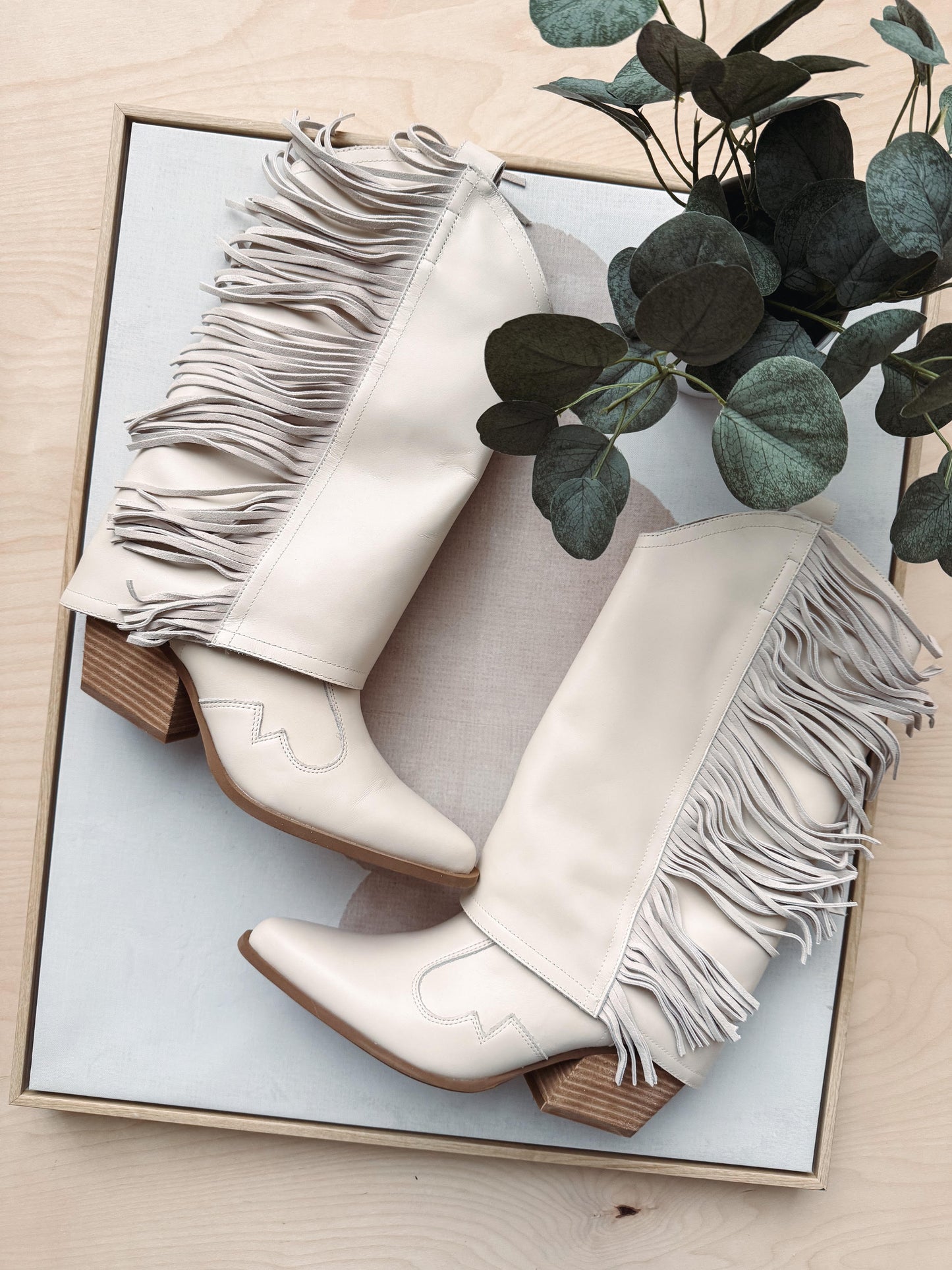 White cowboy boots with fringes on a light wooden surface with greenery.