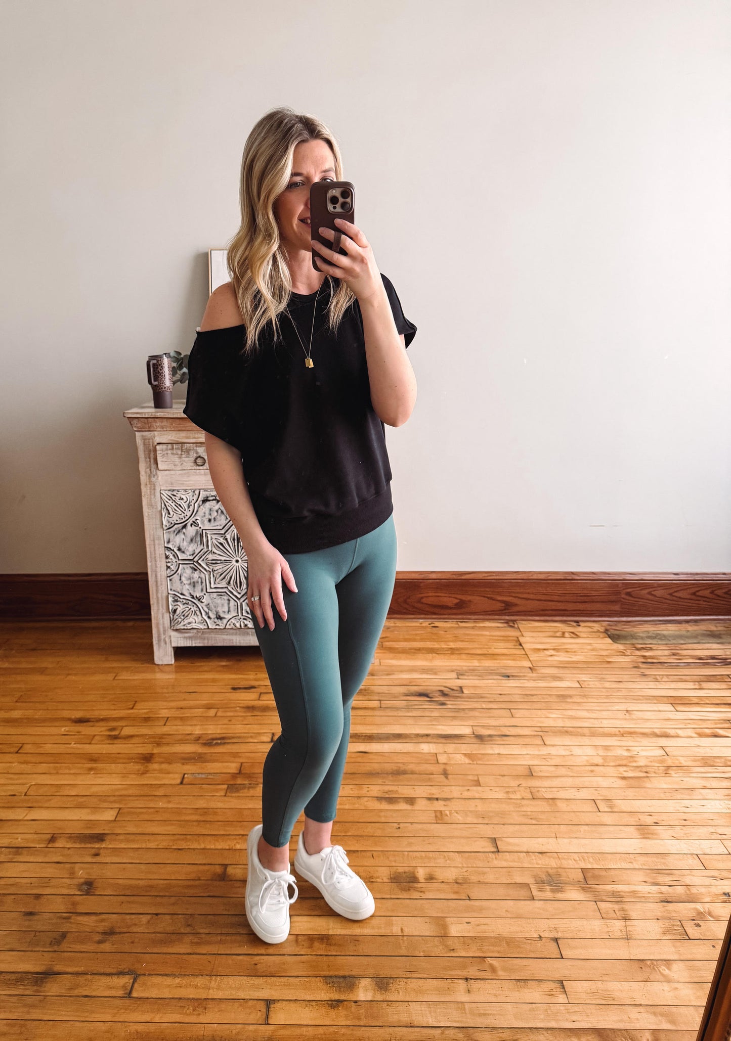 Woman taking a mirror selfie wearing a black top and teal leggings on a wooden floor.