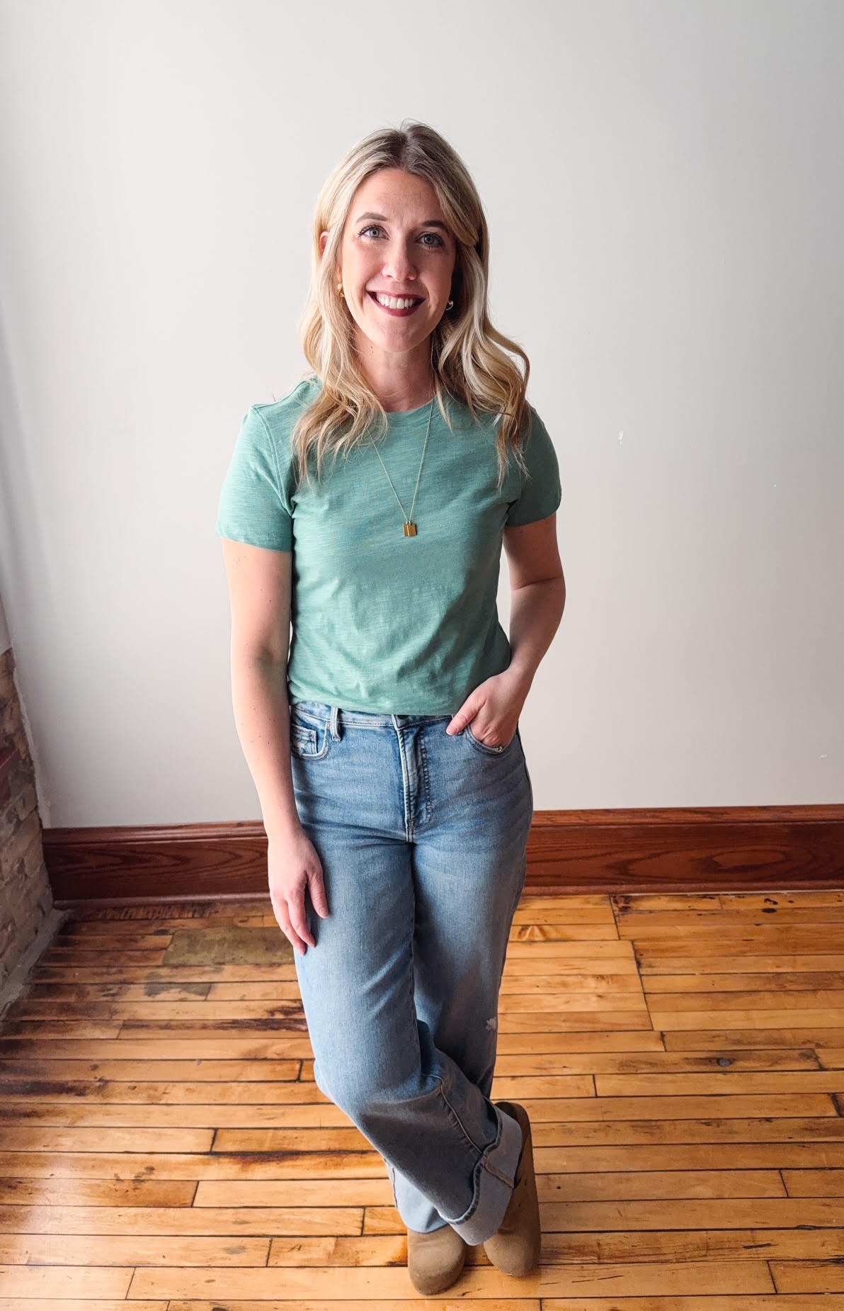 Woman wearing a green t-shirt and blue jeans standing on a wooden floor.