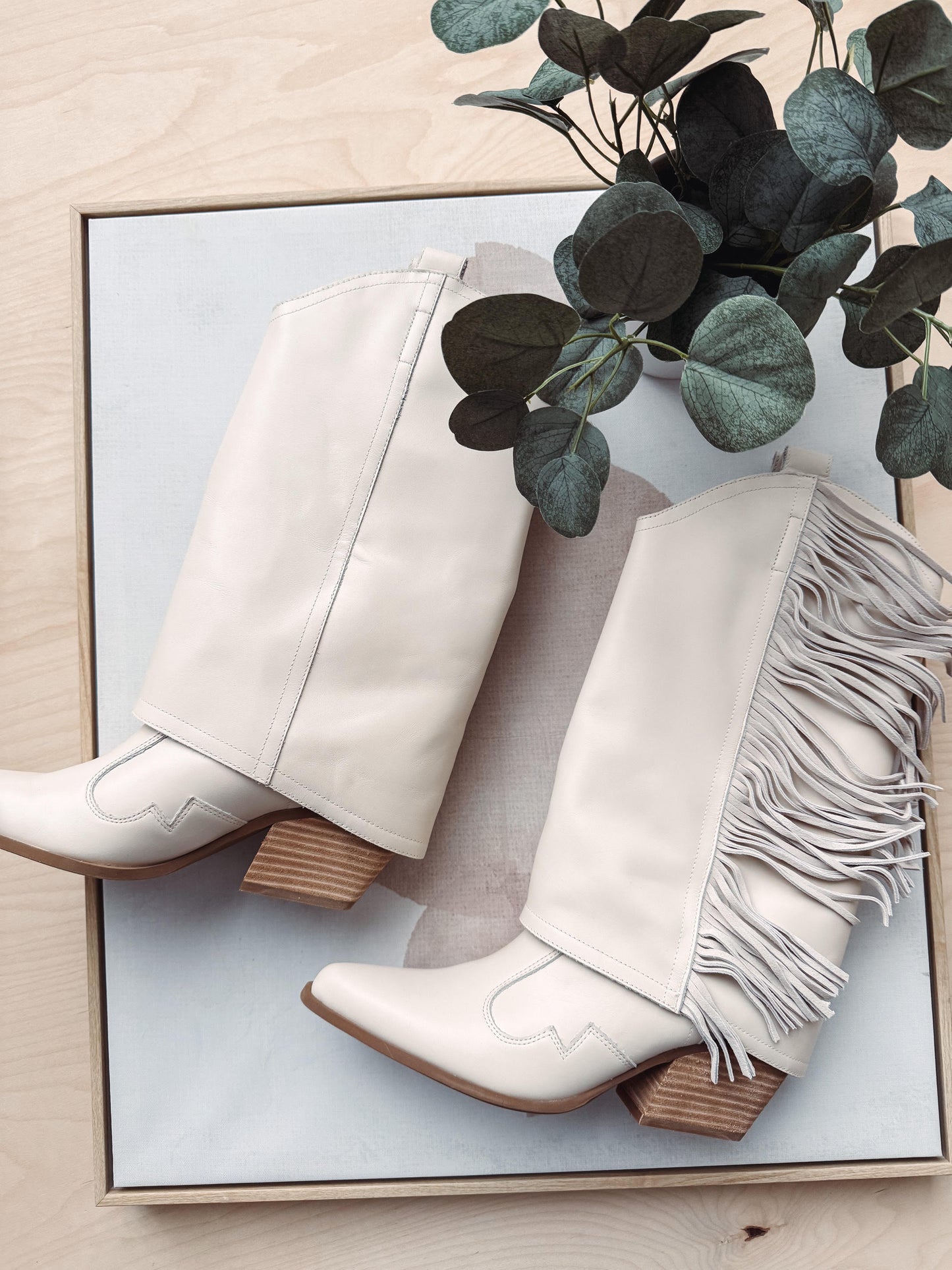 White cowboy boots with fringes on a light wooden surface with greenery.