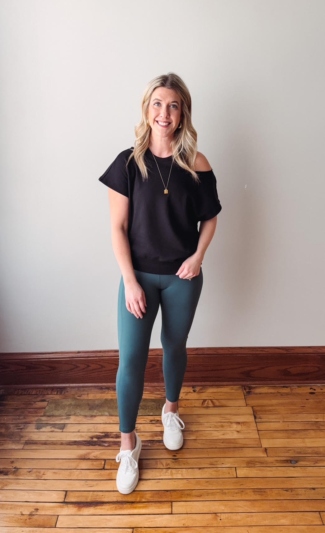 Woman wearing a black top and teal leggings standing on a wooden floor.
