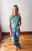Woman wearing a green t-shirt and blue jeans standing on a wooden floor.