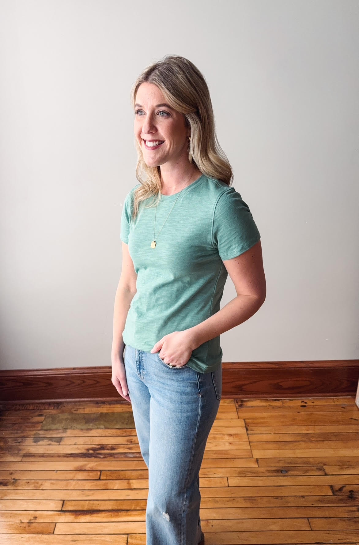 Woman wearing a light green t-shirt and blue jeans standing on a wooden floor.