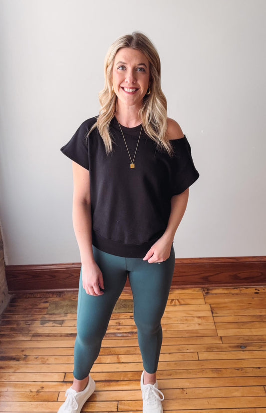 Woman wearing a black off-shoulder top and teal leggings standing on a wooden floor.