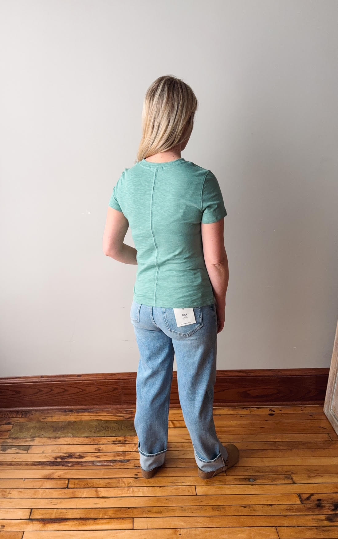 Person wearing a green shirt and blue jeans standing on a wooden floor with a white wall background