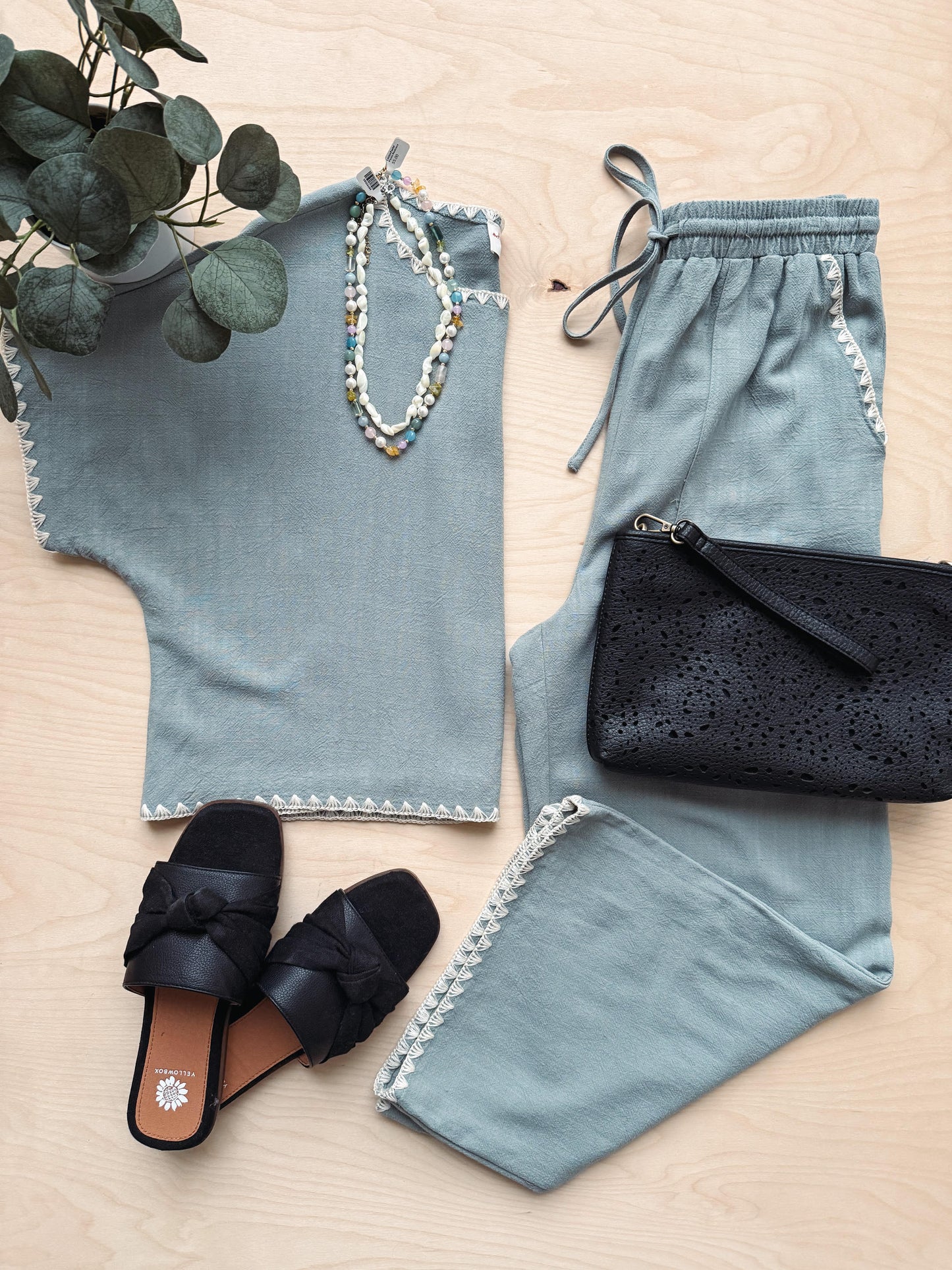 Light blue top, pants, black sandals, and clutch on a wooden surface with greenery.