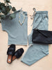 Light blue top, pants, black sandals, and clutch on a wooden surface with greenery.
