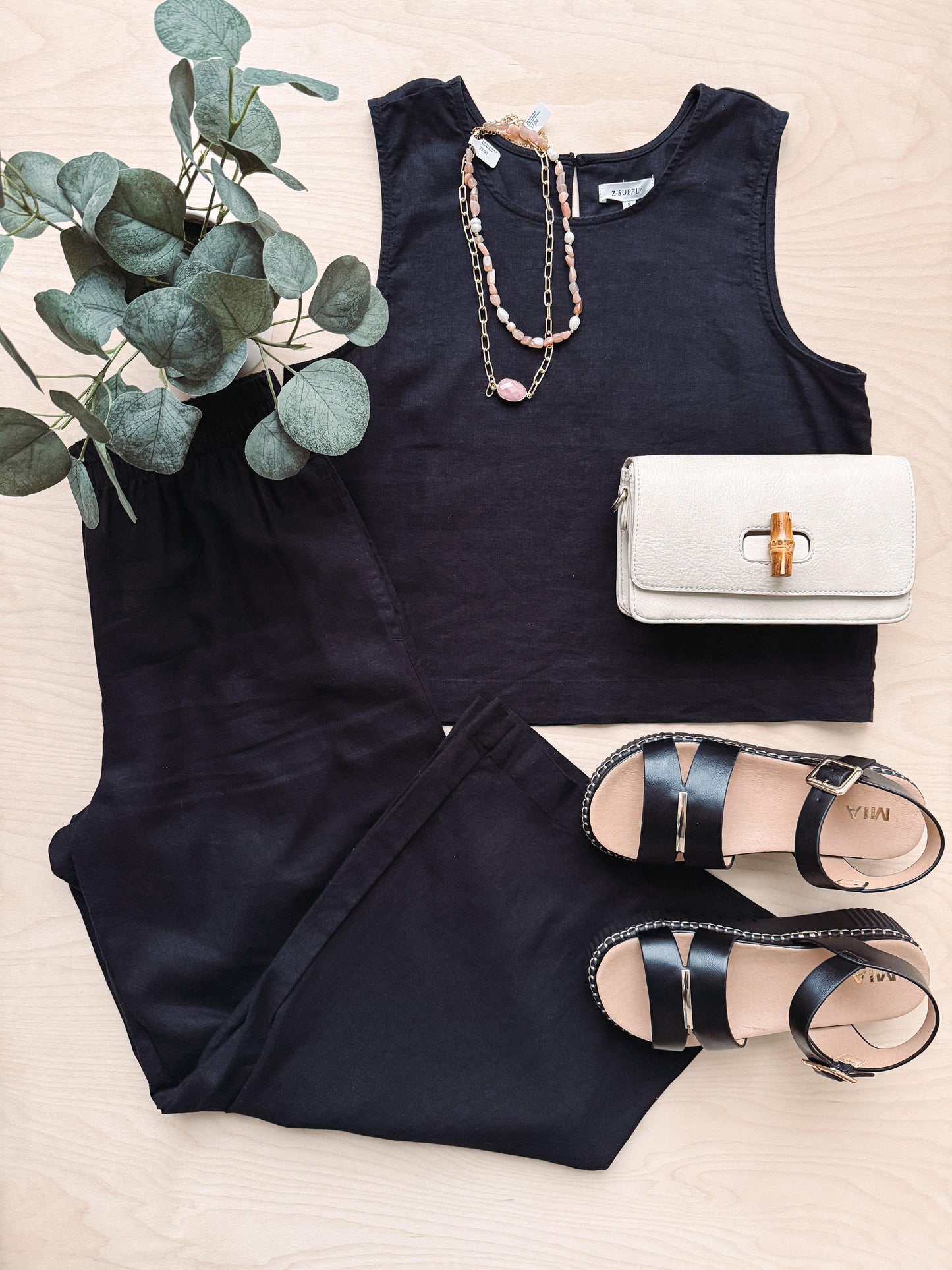 Black sleeveless top, white handbag, and black sandals on a light wooden surface with greenery.
