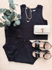 Black sleeveless top, white handbag, and black sandals on a light wooden surface with greenery.