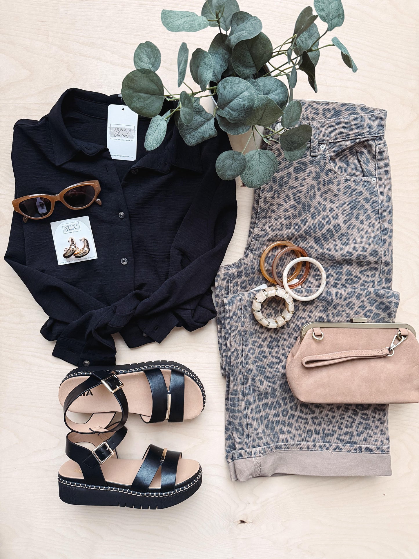 Black shirt, leopard print pants, sandals, beige handbag, sunglasses, and a plant on a light surface.