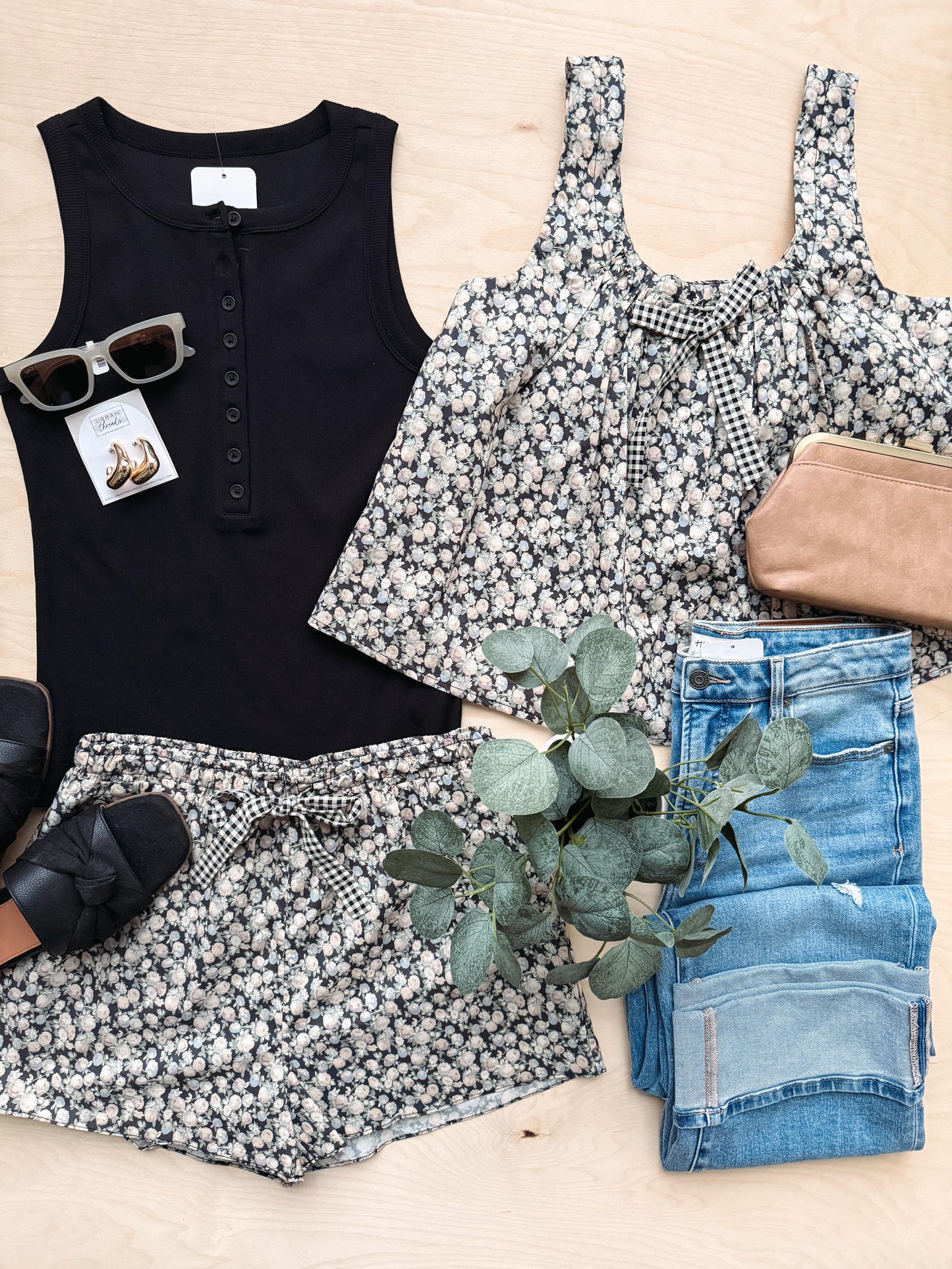 Collection of clothing and accessories including a black tank top, floral shorts, jeans, and sunglasses on a light wooden surface.