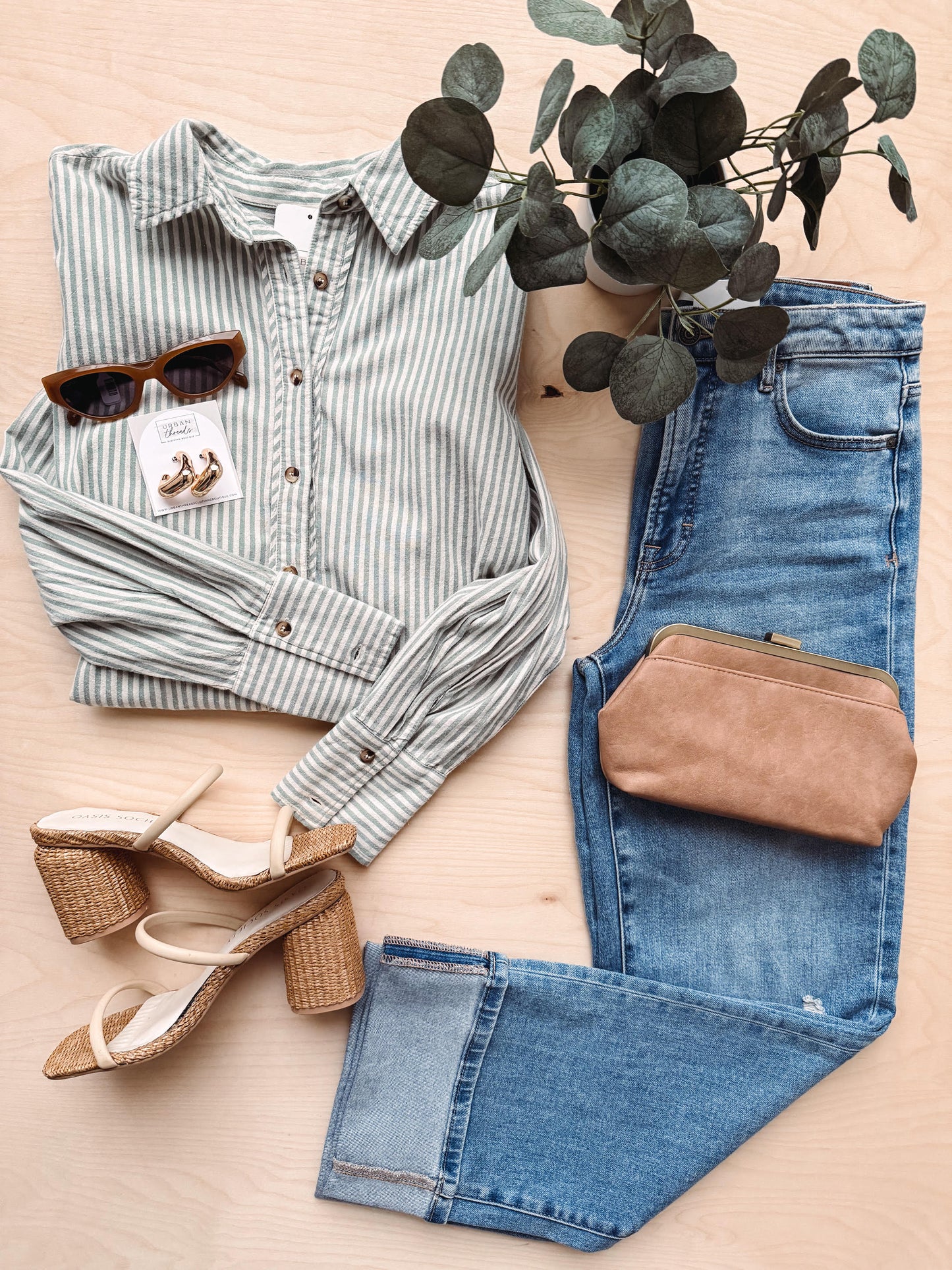 Striped shirt, jeans, sunglasses, and sandals arranged on a light surface with eucalyptus leaves.