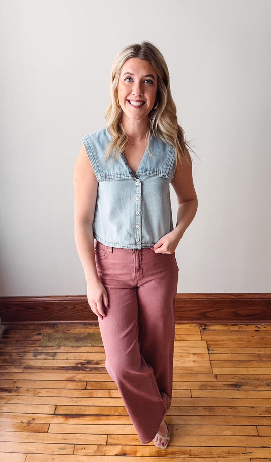 Woman wearing a light blue denim vest over a white shirt and pink pants, standing on a wooden floor.
