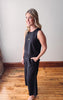 Woman wearing a black sleeveless jumpsuit standing on a wooden floor with a white wall background