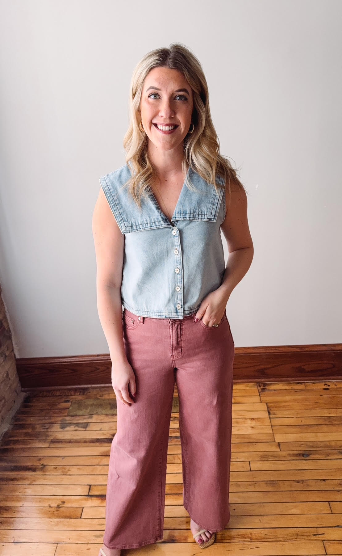 Woman wearing a denim sleeveless shirt and pink pants standing on a wooden floor.