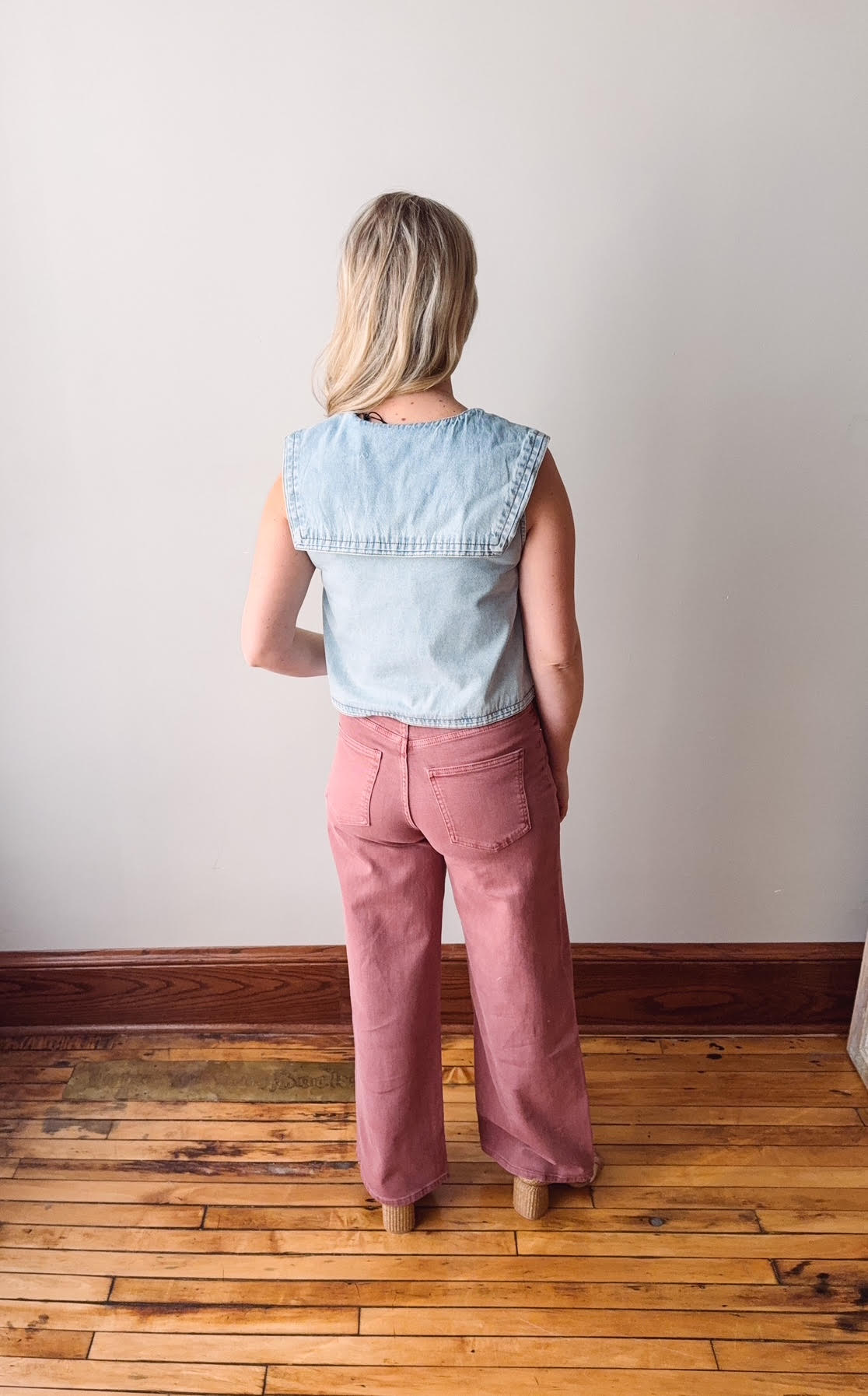 Person wearing a light blue denim top and pink pants standing on a wooden floor with a white wall background
