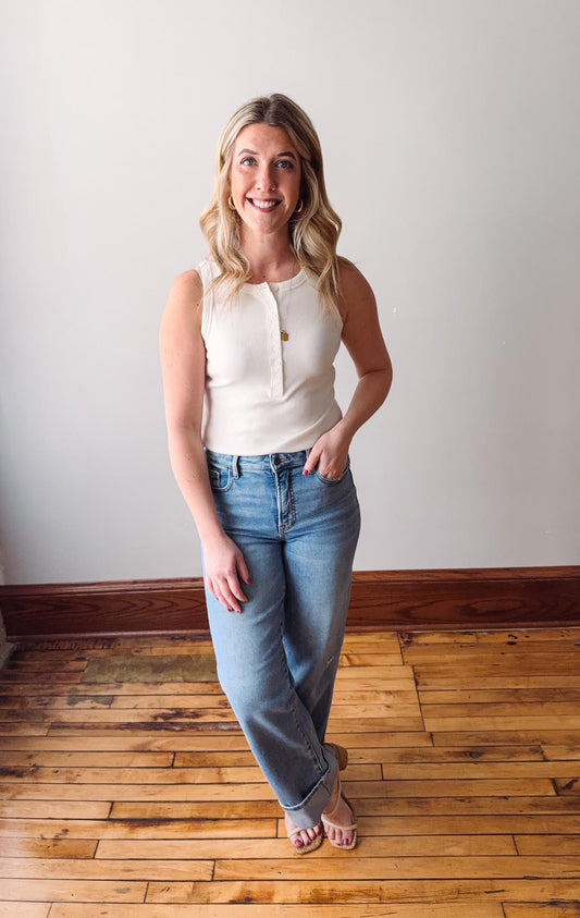 Woman wearing a white sleeveless top and blue jeans standing on a wooden floor.